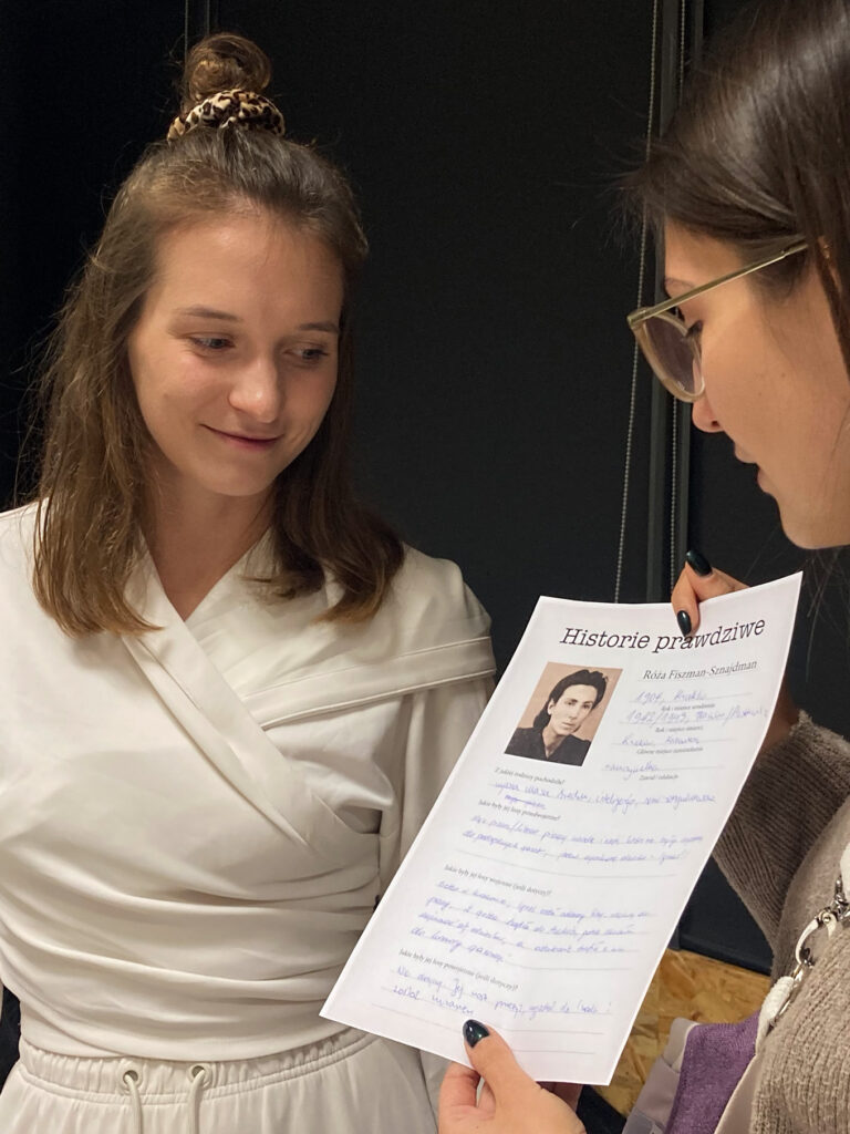 Two people at a workshop, one holding a printed profile sheet titled “True Stories” with handwritten notes and a small portrait photo, engaged in discussion about Polish-Jewish history and perceptions