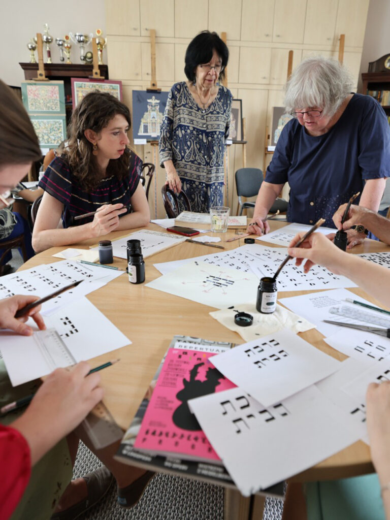 Group of people sitting around a table at a Hebrew calligraphy workshop, practicing writing Hebrew letters with ink and brushes