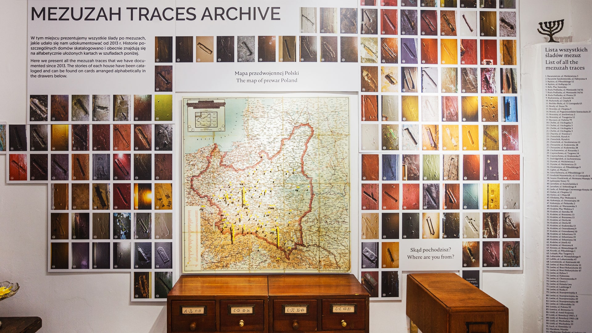 Mezuzah museum display titled “Mezuzah Traces Archive,” showing a wall filled with photographs of mezuzah traces from various homes, a map of prewar Poland marked with locations, and drawers below for catalogued stories and documents