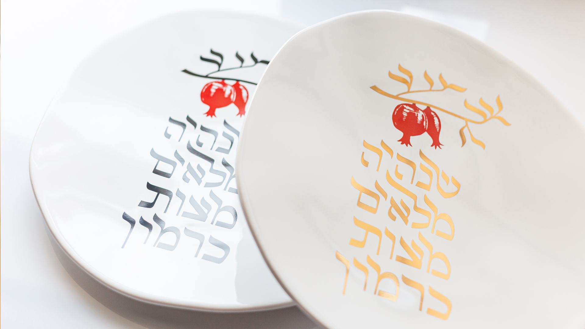 Two decorative plates for Rosh HaShana, featuring pomegranate and Hebrew calligraphy designs with subtle reference to mezuzah trace, designed by MI POLIN design