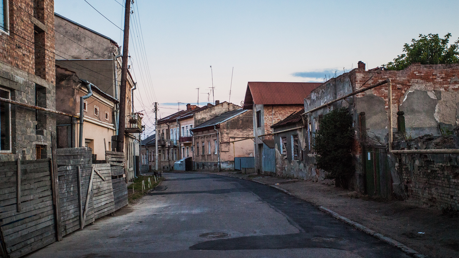 Quiet street lined with old houses and weathered fences in a small Eastern European town at dusk, linked by mezuzah trace